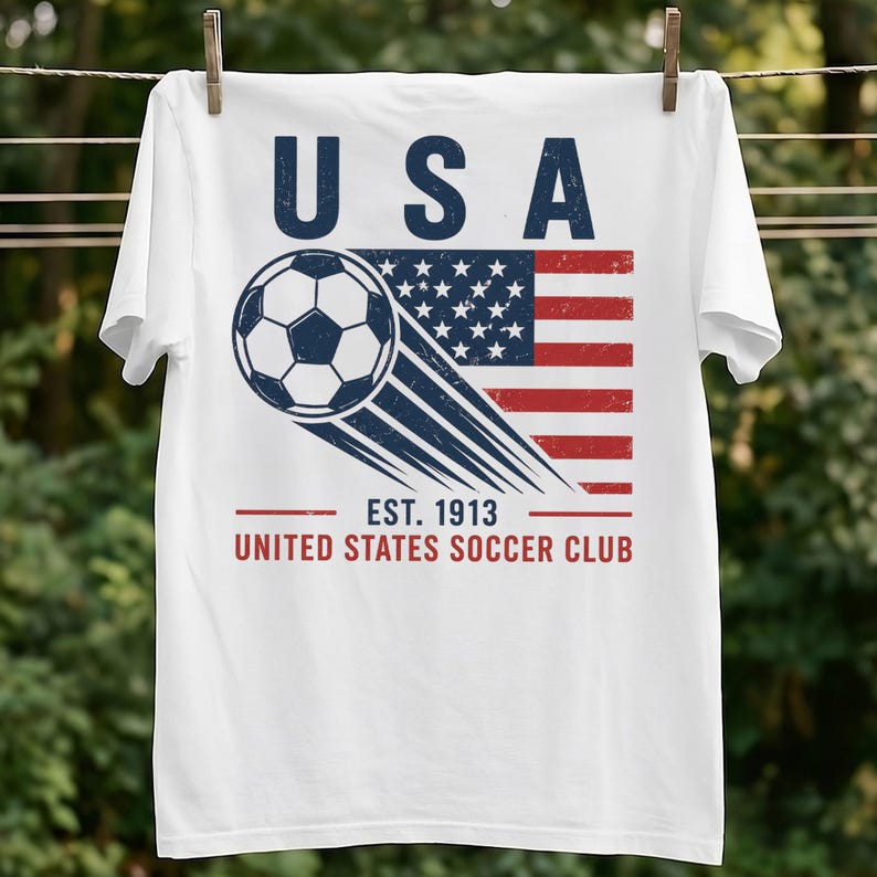Puede incluir: Una camiseta blanca con un gr&aacute;fico de un bal&oacute;n de f&uacute;tbol en movimiento sobre una bandera estadounidense desgastada. El texto USA aparece en la parte superior, y EST. 1913 UNITED STATES SOCCER CLUB en la parte inferior en letras azul oscuro y rojo.