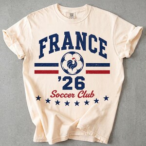 Op de afbeelding: Een cr&egrave;mekleurig T-shirt met korte mouwen en een verweerde opdruk met de tekst FRANCE, een voetbal met een haan-icoon, het getal 26 en de woorden Soccer Club, geaccentueerd door horizontale blauwe en rode strepen en een rij blauwe sterren.