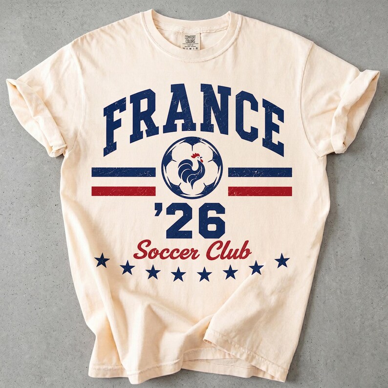 Op de afbeelding: Een cr&egrave;mekleurig T-shirt met korte mouwen en een verweerde opdruk met de tekst FRANCE, een voetbal met een haan-icoon, het getal 26 en de woorden Soccer Club, geaccentueerd door horizontale blauwe en rode strepen en een rij blauwe sterren.