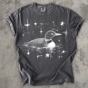 May include: A charcoal gray short-sleeve t-shirt featuring a white line-art illustration of a loon swimming on water. The design is surrounded by small, star-like sparkles. The shirt has a crew neckline and a visible brand tag at the inner collar.