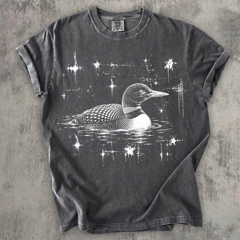 May include: A charcoal gray short-sleeve t-shirt featuring a white line-art illustration of a loon swimming on water. The design is surrounded by small, star-like sparkles. The shirt has a crew neckline and a visible brand tag at the inner collar.