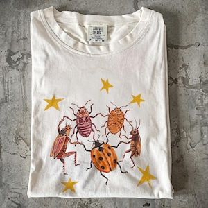 May include: A white short-sleeve t-shirt featuring a graphic of five insects holding hands in a circle, surrounded by yellow stars. The insects include a ladybug, beetles, and a cricket. The inner neck label reads Comfort Colors size M.