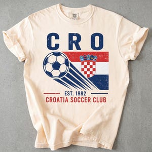 May include: A cream-colored short-sleeve t-shirt featuring a graphic with the text CRO, a soccer ball, the Croatian flag, and the text EST. 1992 CROATIA SOCCER CLUB. The design has a distressed, vintage-inspired texture in blue, red, and white.