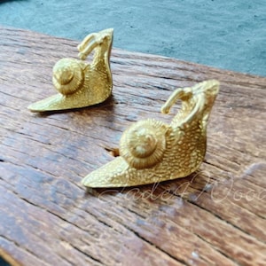 May include: Two gold snail sculptures on a wooden surface. The snails are detailed and have a textured finish.