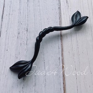 Puede incluir: Tirador de armario negro con un diseño de hoja decorativa en cada extremo. El tirador tiene un centro retorcido en forma de cuerda. El herraje está sobre un fondo de madera blanca y turquesa.