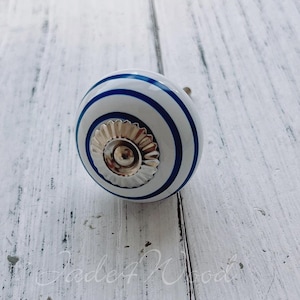 May include: A white ceramic cabinet knob with blue stripes and a silver-coloured metal base. The knob is round and has a decorative design. The background is a white wooden surface.