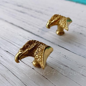 Pode incluir: Dois botões de armário de cabeça de águia dourados com padrões de penas detalhados.