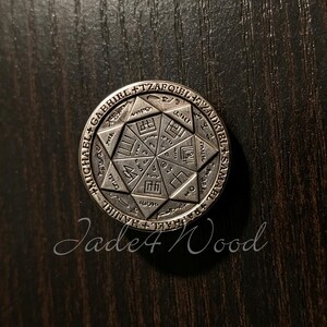 May include: A circular silver-toned metal coin featuring an engraved eight-pointed star design. The outer rim displays the names Michael, Gabriel, Tzafqiel, Tzadkiel, Samael, Raphael, and Haniel. The centre contains geometric symbols and lines.