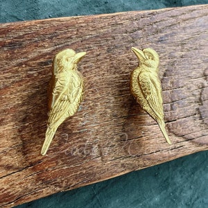 May include: Two gold bird-shaped wall hooks mounted on a wooden surface. The hooks are detailed with feathers and have a realistic bird design.