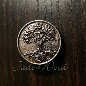 Puede incluir: Un botón metálico circular de tono plateado con un diseño en relieve detallado de un árbol con ramas extendidas y raíces visibles. El texto Jade4Wood está impreso en letra cursiva debajo del botón sobre la superficie de madera oscura.