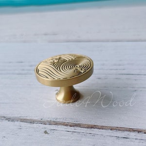 Pode incluir: Um botão de armário dourado com uma base circular e um design de ondas. O botão está sobre uma superfície de madeira branca.