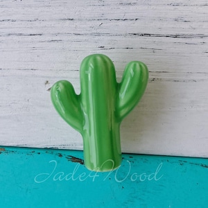 Può includere: Una statuetta di cactus in ceramica verde con due braccia. Il cactus è su uno sfondo di legno bianco e blu.