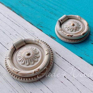 Könnte beinhalten: Zwei weiße und goldene dekorative Schubladenknöpfe mit einem Ringgriff. Die Knöpfe haben ein florales Design und befinden sich auf einer weißen und blauen Holzoberfläche.