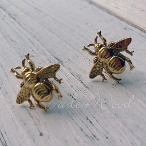 Puede incluir: Dos pendientes dorados con forma de abeja. Los pendientes presentan alas, antenas y segmentos corporales detallados. Se presentan sobre una superficie clara y texturizada. Son una pieza de joyería.
