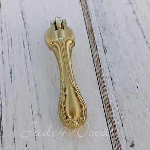 May include: Gold-toned metal cabinet pull with an ornate design. The pull has a long, slender handle with a decorative floral pattern.