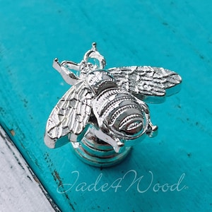 Può includere: Pomo per armadio a forma di ape argentato con un designo dettagliato. Il pomo è su uno sfondo blu turchese.