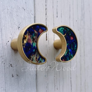 May include: Two crescent-shaped cabinet knobs with a gold finish and a colorful, marbled inlay of blues, greens, and other hues. The knobs are mounted on a white wooden surface.