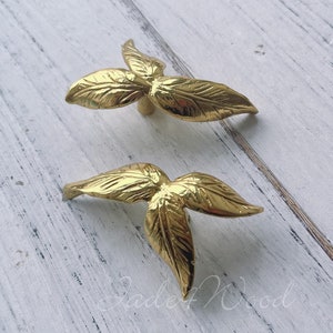 May include: Two gold leaf-shaped cabinet knobs with a simple design. The knobs are made of metal and have a smooth, polished finish.