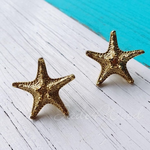Könnte beinhalten: Ein Paar goldfarbene Seestern-Ohrringe. Die Ohrringe sind aus Metall und haben eine strukturierte Oberfläche.