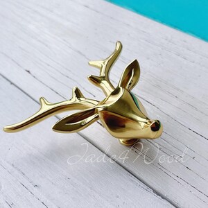 Puede incluir: Pomo de armario de cabeza de ciervo dorado con un diseño detallado. El pomo está sobre una superficie de madera blanca.