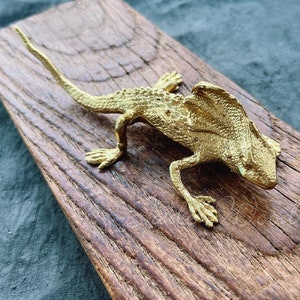 Könnte beinhalten: Eine goldfarbene Metallfigur eines Leguan mit einem Kragen um den Hals. Der Leguan ist auf einer Holzoberfläche platziert.