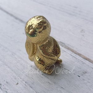 Pode incluir: Uma figurinha de coelho dourada com uma superfície detalhada e texturizada. O coelho está sentado com a cabeça inclinada para baixo e as patas enfiadas.