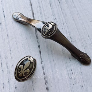 May include: Two antique brass cabinet pulls with a fleur-de-lis design. The pulls are oval shaped with a raised design and a textured finish.