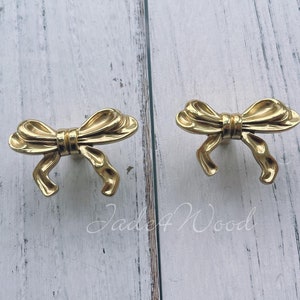 Peut inclure: Deux boutons de cabinet en forme de nœud de couleur or. Les boutons sont en métal et ont une finition lisse et polie.