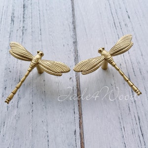 Puede incluir: Dos tiradores de cajón de libélula dorados. El diseño de la libélula es detallado y realista. Los tiradores están montados en una superficie de madera blanca.