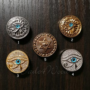 May include: Five circular metal buttons labelled 1 through 5. Buttons 1, 2, 3, and 4 feature the Eye of Horus with a blue centre. Button 1 is silver, 2 is gold, 3 is gold with rhinestones, and 4 is silver with rhinestones. Button 5 is bronze with a star.