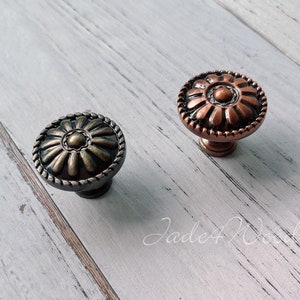 May include: Two ornate cabinet knobs with a floral design. The knob on the left is a dark bronze color, and the knob on the right is a copper color.