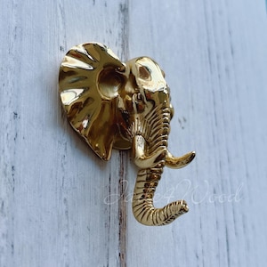 May include: Gold-coloured elephant-shaped hook mounted on a white wooden surface. The hook is a decorative accent for a home or office.