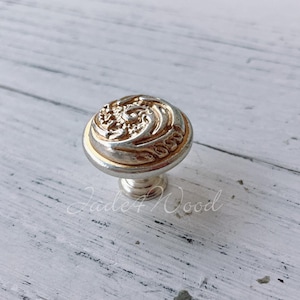 Może przedstawiać: Dekoracyjny gałka do szafki w kolorze srebrnym i złotym z kwiatowym wzorem.