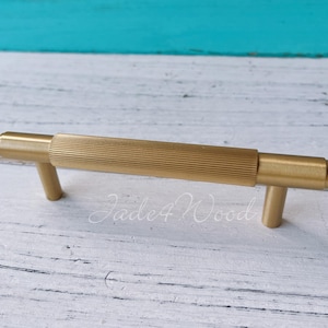 Pode incluir: Um puxador de armário dourado com uma seção central canelada e dois postes cilíndricos. O hardware é mostrado em um fundo branco e turquesa. O texto "Jade4Wood" é visível.