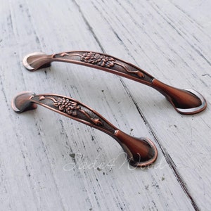 Könnte beinhalten: Zwei verzierte Kupfer-Schubladengriffe mit floralem Design. Die Griffe befinden sich auf einer weißen Holzoberfläche.