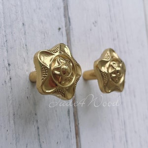 Peut inclure: Deux boutons de porte en métal doré avec un motif floral. Les boutons sont fixés à une surface en bois blanc.