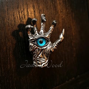 May include: A silver-toned metal cabinet knob shaped like a hand with elongated fingers and sharp nails. The palm features a prominent, bright blue eye with a black pupil. The surface of the hand has a textured, organic appearance. The text Jade & Wood is visible.