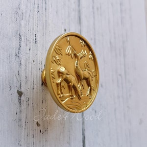 May include: A gold-colored cabinet knob with a detailed relief of two deer in a forest setting. The knob is round with a small stem for attachment. The background is a white, distressed wood surface.