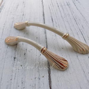 Puede incluir: Dos tiradores de armario de cerámica de color crema con detalles dorados. Los tiradores tienen una forma curva y un diseño decorativo en los extremos.