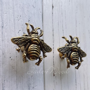 May include: Two bronze-colored bee-shaped cabinet knobs. The knobs are detailed with wings, legs, and body segments. They are set against a white, weathered wood background. The knobs are likely for furniture or decorative use.