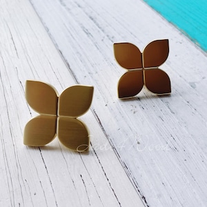 May include: Two gold-toned metal cabinet knobs shaped like four-leaf clovers. The knobs are on a white wooden surface.