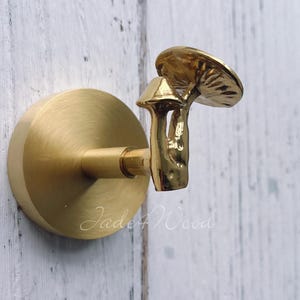 Puede incluir: Un gancho en forma de hongo de latón montado sobre una base redonda. El gancho presenta un diseño detallado de hongo con un sombrero y tallo texturizados. Está fijado a una superficie de madera blanca.