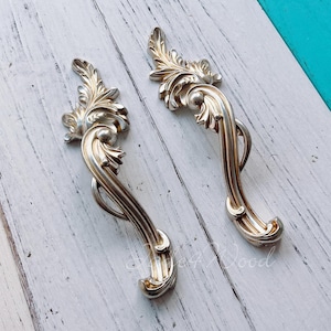 May include: Two ornate silver-toned drawer pulls with a floral design. The pulls are shaped like a curved S with a decorative flourish at the top.