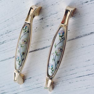 May include: Two gold cabinet pulls with a white ceramic insert featuring a floral design. The pulls are oval shaped and have a smooth, polished finish.