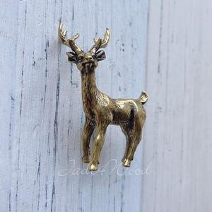 Puede incluir: Una detallada figura de ciervo en color latón con cornamenta. El ciervo está sobre una superficie blanca y azul claro. El texto "Jade4Wood" es visible en la superficie. Es un objeto decorativo.