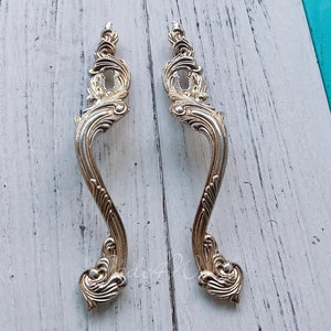 May include: Two ornate silver-toned cabinet door pulls with a decorative, swirling design.