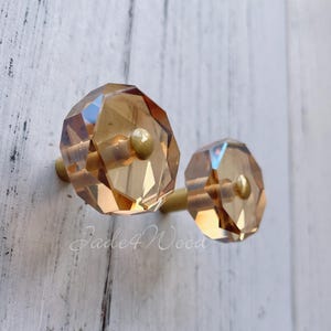 以下が含まれることがあります： 琥珀色のクリスタルキャビネットノブ2つ。ゴールドトーンの金具付き。ノブは多面的な幾何学模様で、白い木製の表面に取り付けられています。「Jade4Wood」の文字が画像に表示されています。