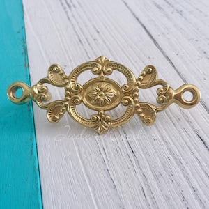 Pode incluir: Puxador de gaveta de metal dourado com um design ornamentado. O puxador tem um motivo floral no centro e uma borda decorativa.