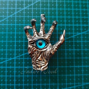 May include: A silver-toned metal pendant shaped like a hand with elongated, claw-like fingers. The palm features a prominent, realistic blue eye with a dark pupil centered in the middle. The surface of the hand has a textured, weathered appearance.