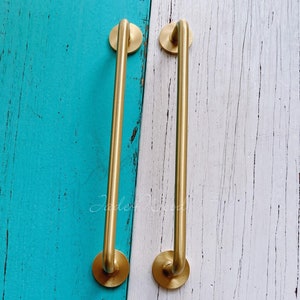 Pode incluir: Duas puxadores de armário de metal dourado com bordas arredondadas e um acabamento liso. Os puxadores estão montados sobre um fundo de madeira azul e branco.